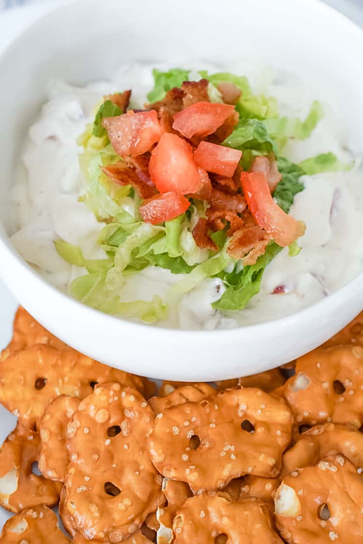 a bowl of blt dip topped with shredded lettuce, diced tomatoes and bacon, served with pretzel crisps