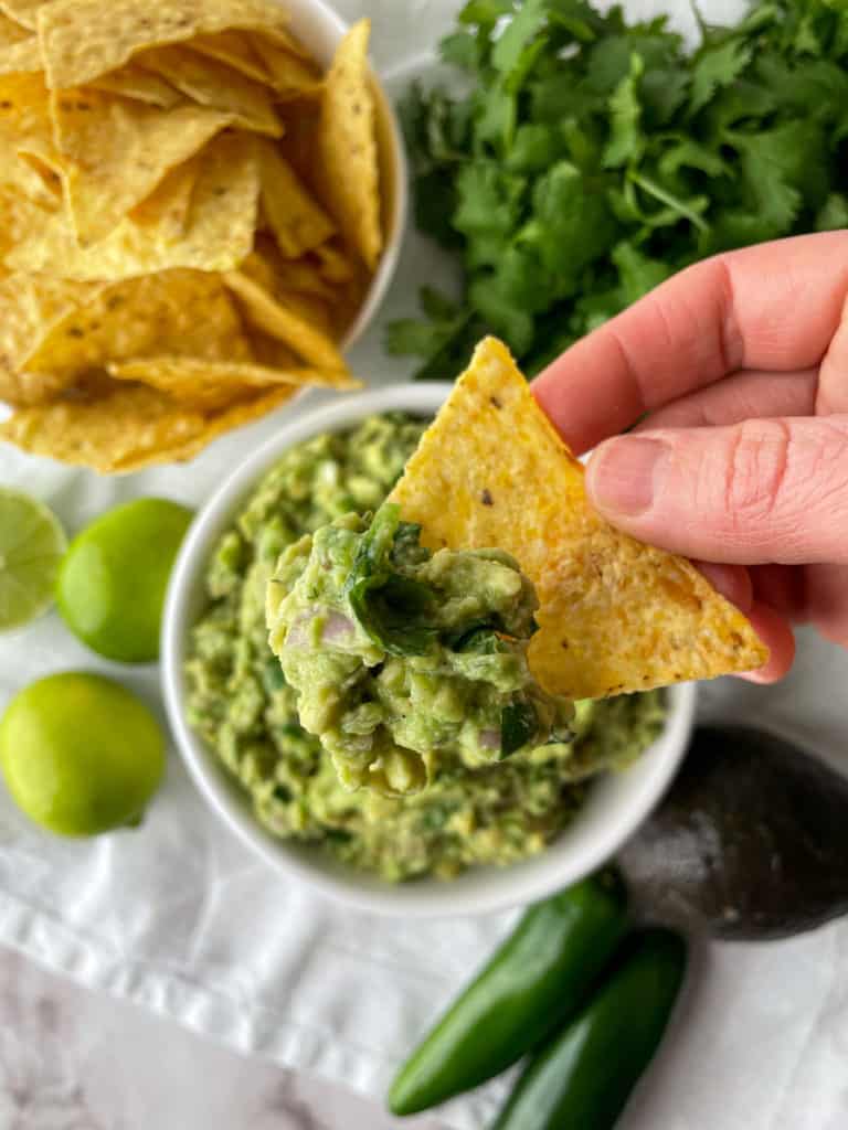 tortilla chip being dipped in guacamole without tomatoes with lime and other guac ingredients on the side