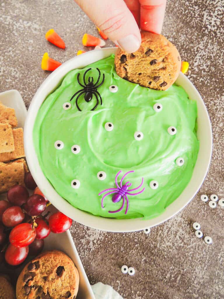 chocolate chip cookie being dipped in green Halloween dessert dip with edible candy eyes