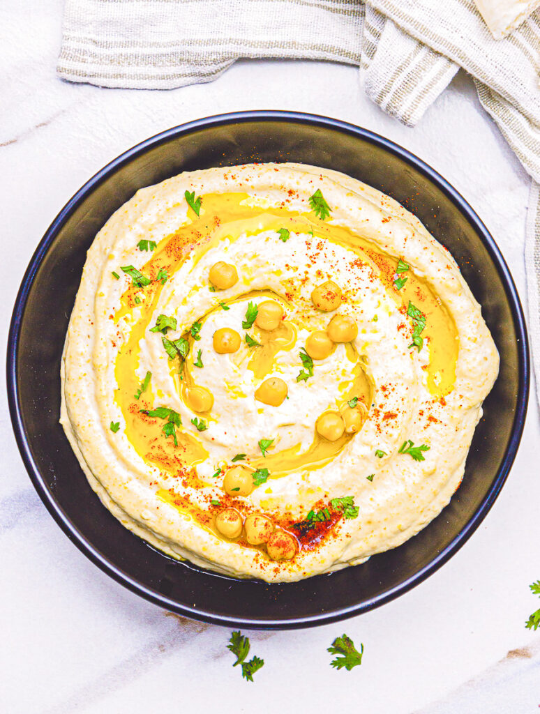 Authentic Hummus Recipe made with chickpeas, tahini and spices in a black bowl garnished with olive oil, paprika and chickpeas