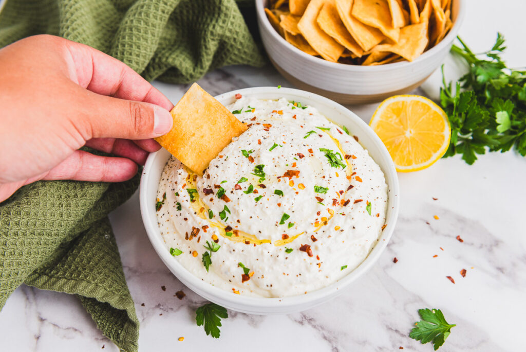 pita chip being dipped in whipped feta in a white bowl with a side of chips