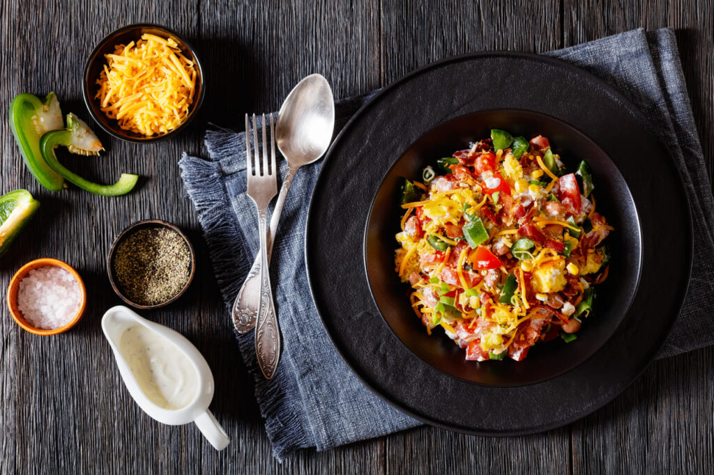 pinto beans bacon cornbread salad with tomatoes, peppers, corn and cheddar cheese in black bowl on dark wood table, horizontal view from above, flat lay
