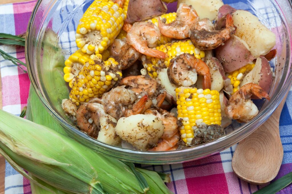 A glass bowl is filled with corn on the cob, red potatoes, and shrimp - a classic shrimp boil Everything is seasoned to perfection and steaming hot You can even see the condensation on the side of the bowl from the steam The bowl rests atop a plaid