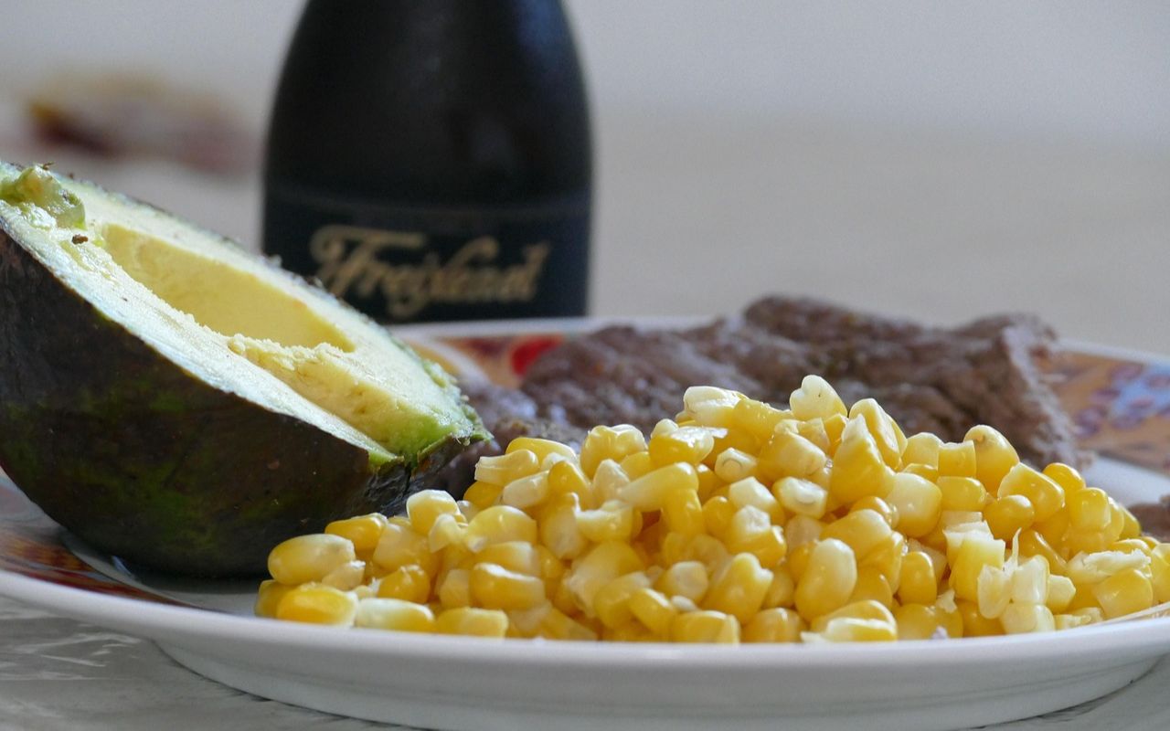 Avocado, corn, and steak on a plate with a bottle in the background