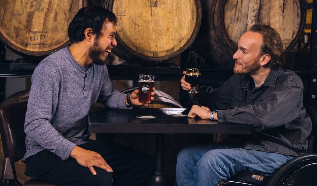 Two Men Sitting at the Table Holding Drinking Glasses While Smiling