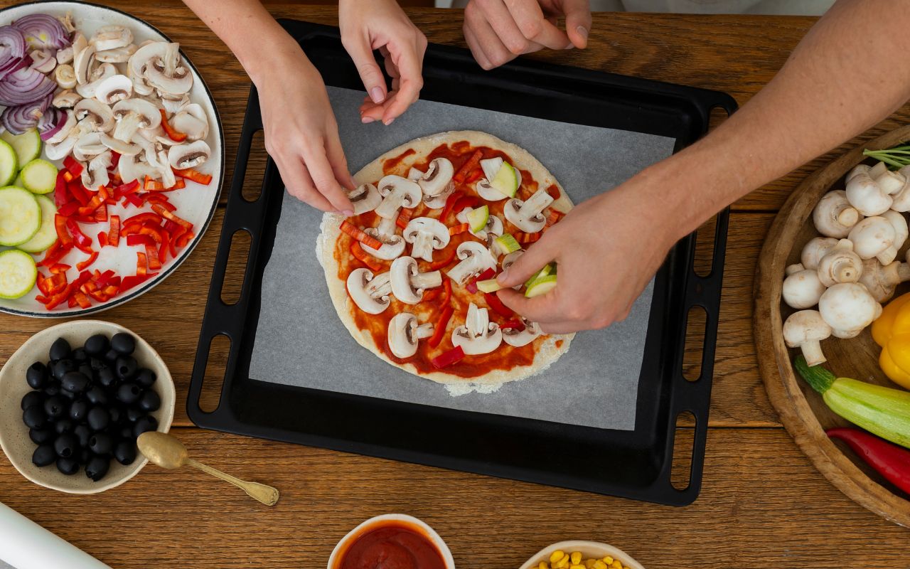 Hands adding veggies to homemade pizza on a parchment-lined tray