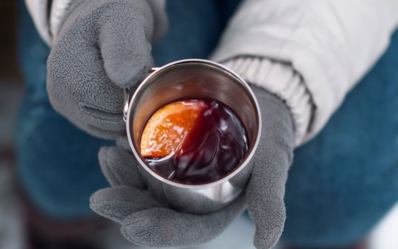 Gloved hands holding a metal mug with a dark drink and an orange slice