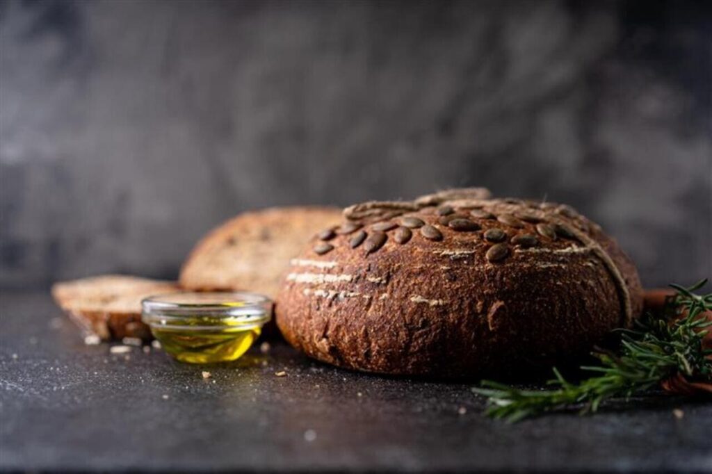 Sliced sourdough bread from whole grain flour and pumpkin seeds on a grid, olive oil and black olive on a rustic wooden table.