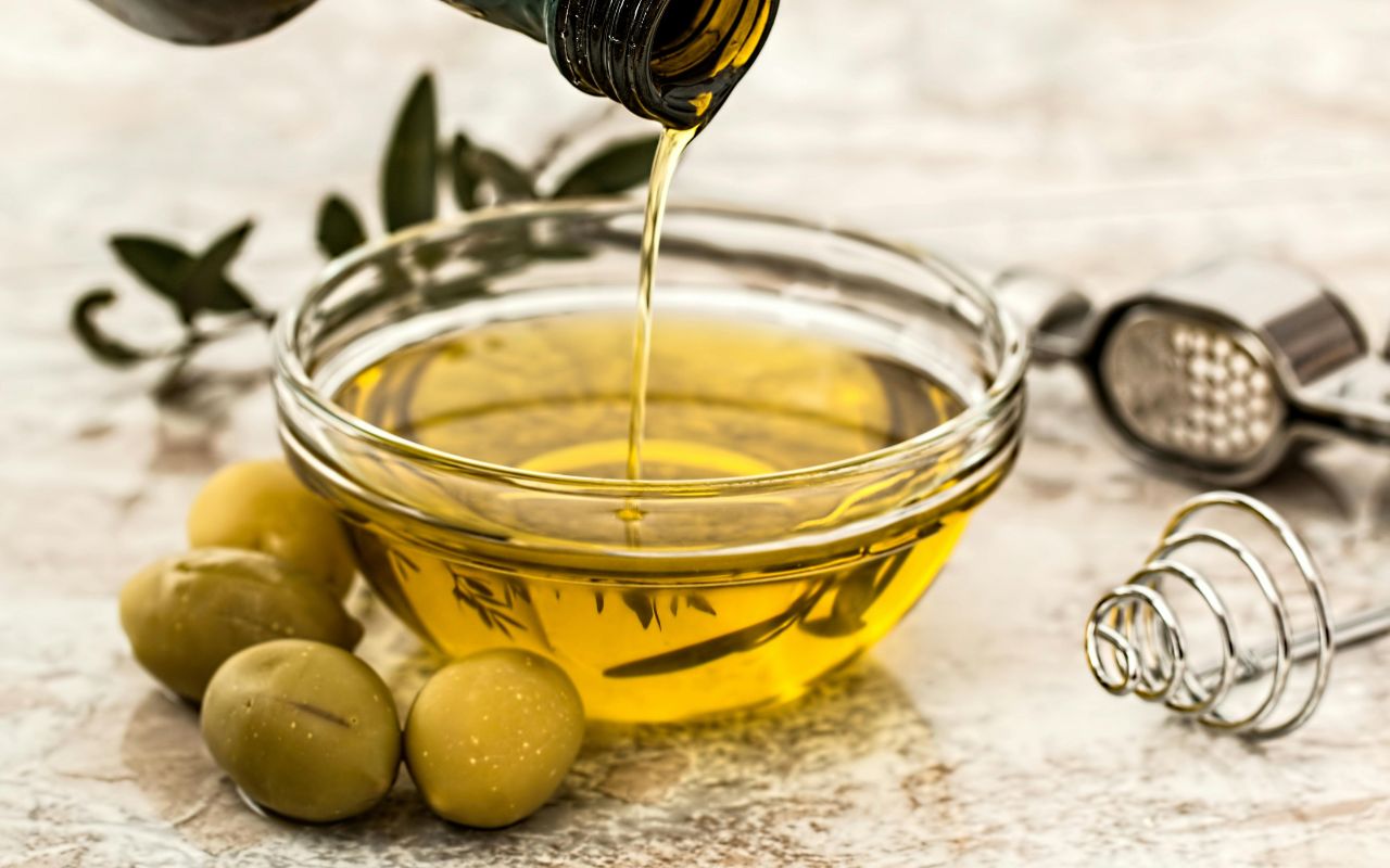 Olive oil pouring into a glass bowl beside green olives