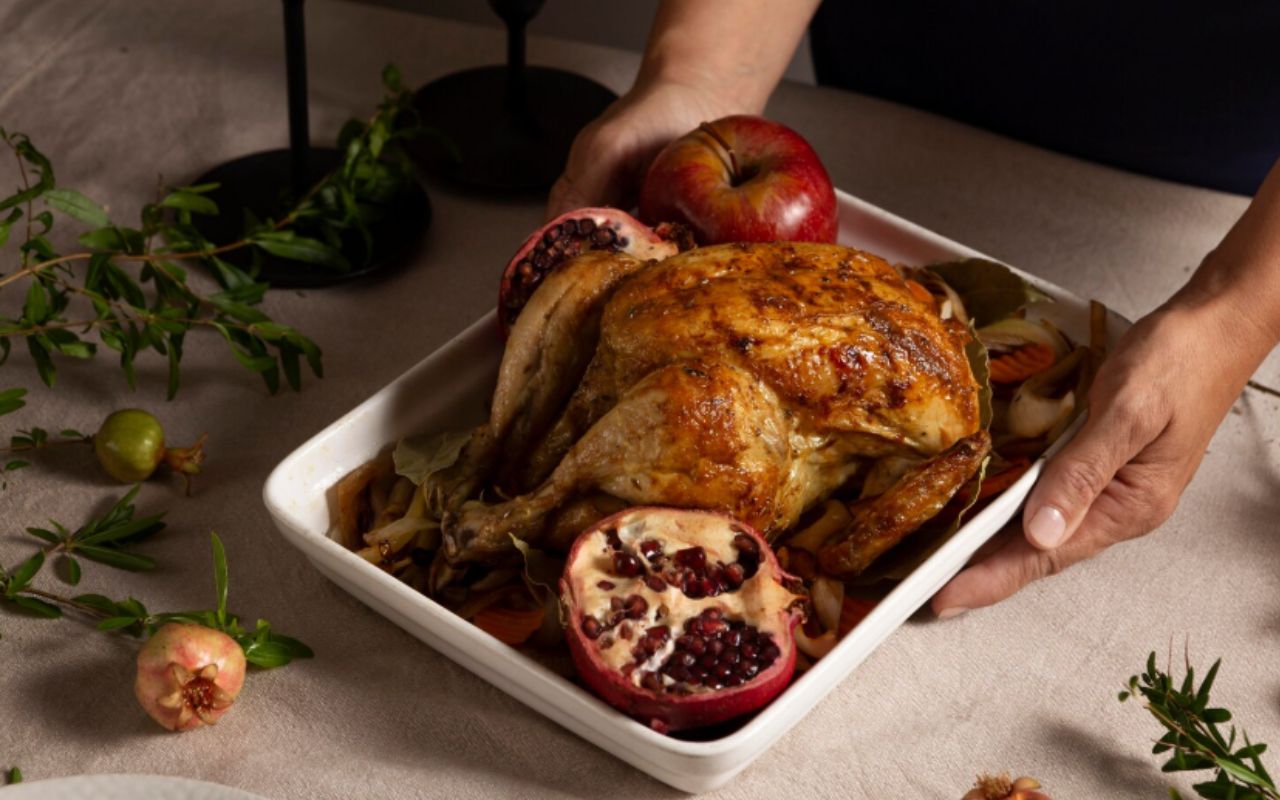 Hand holding a roasted chicken surrounded by apples, pomegranate, and herbs
