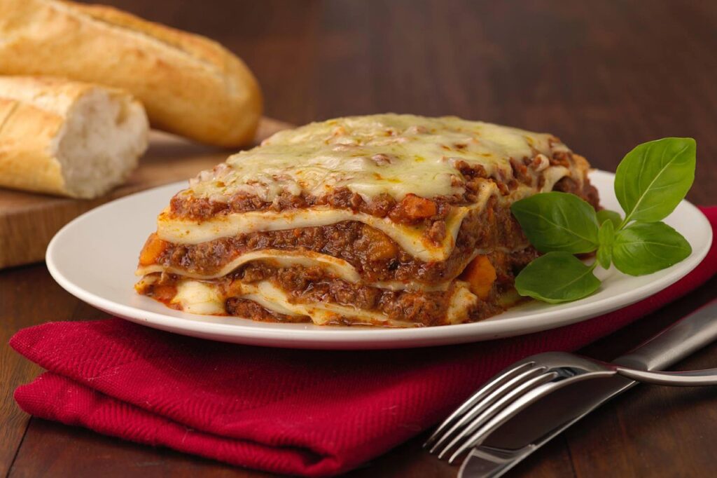 A slice of lasagna on a white plate with melted cheese stretching.