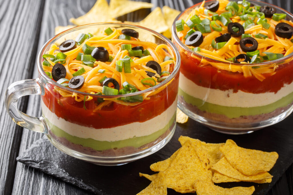 A glass baking dish showing colorful layers of seven-layer dip with chips around it.