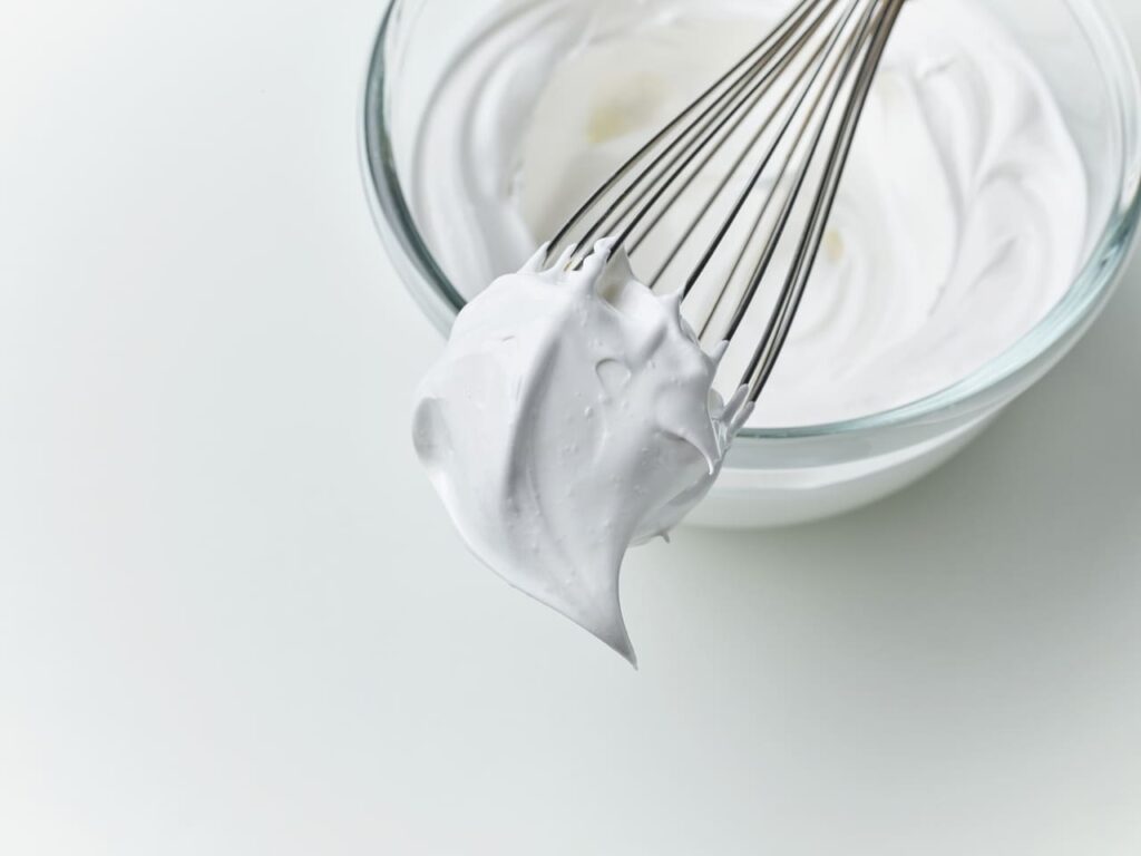 glass bowl of whipped egg whites cream on white kitchen table background, top view
