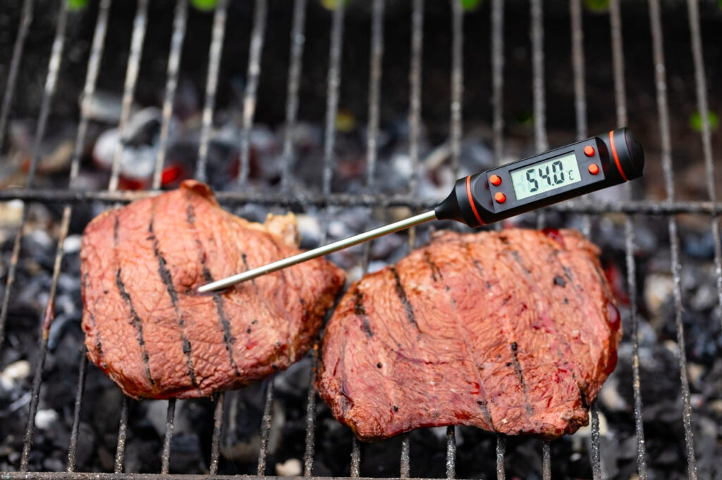 Close-up shot of digital meat thermometer inside grilled steak on barbecue showing the right temperature for Medium Rare doneness