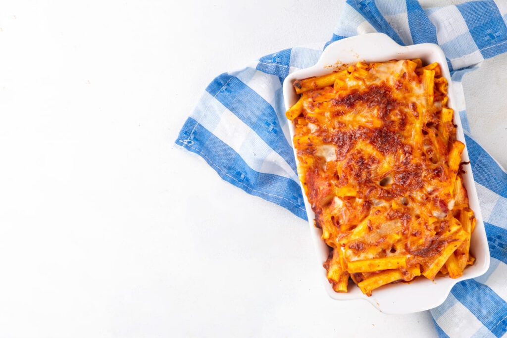 Baked Ziti pasta with bolognese or marinara tomato sauce and cheese, in baked pan and plate, on white kitchen table copy space