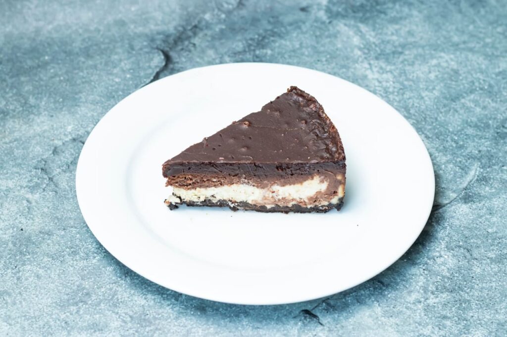 A deliciously rich and creamy nutella oreo cheesecake slice is placed elegantly on a while plate with a minimalistic background.