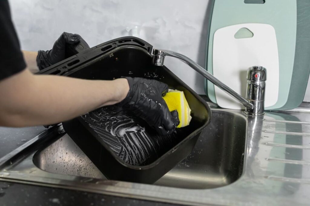 A person's gloved hands diligently scrub a black air fryer basket with a yellow sponge in a stainless steel kitchen sink, highlighting domestic cleanliness