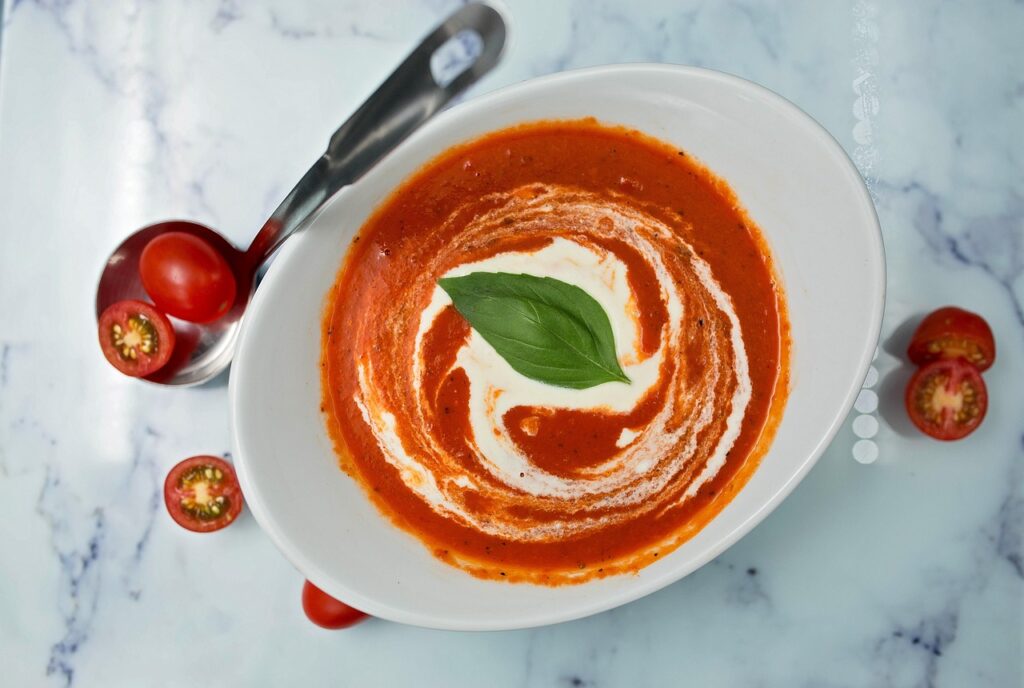 Bright red tomato basil soup in a white bowl with a grilled cheese sandwich.